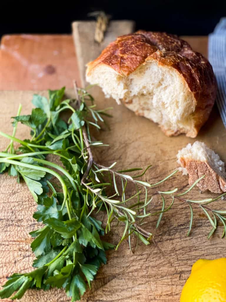 Bread board with fresh herbs, crusty bread, and lemon. To display ingredients used in the chicken noodle soup recipe.