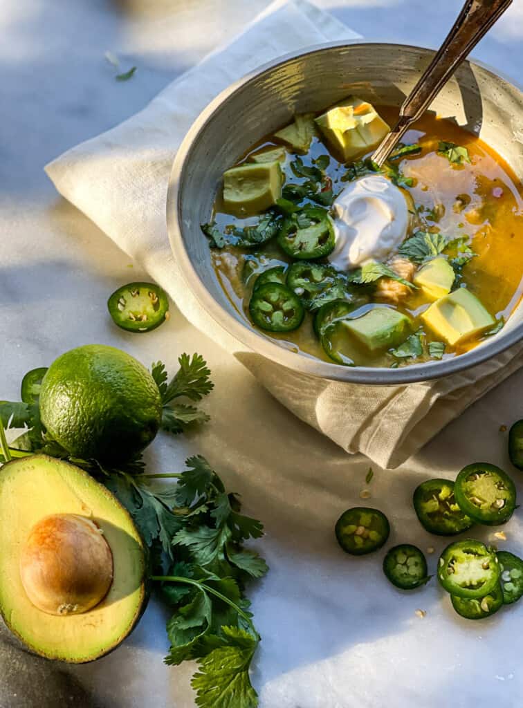A warming bowl of white chicken chili topped with avocado chunks, sour cream, jalapeno slices, and cilantro leaves.