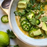 A warming bowl of white chicken chili topped with avocado chunks, sour cream, jalapeno slices, and cilantro leaves. A hand is seen squeezing a lime into the bowl.