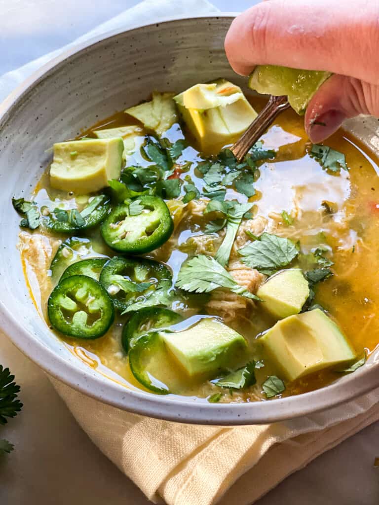 A warming bowl of white chicken chili topped with avocado chunks, sour cream, jalapeno slices, and cilantro leaves. A hand is seen squeezing a lime into the bowl. 