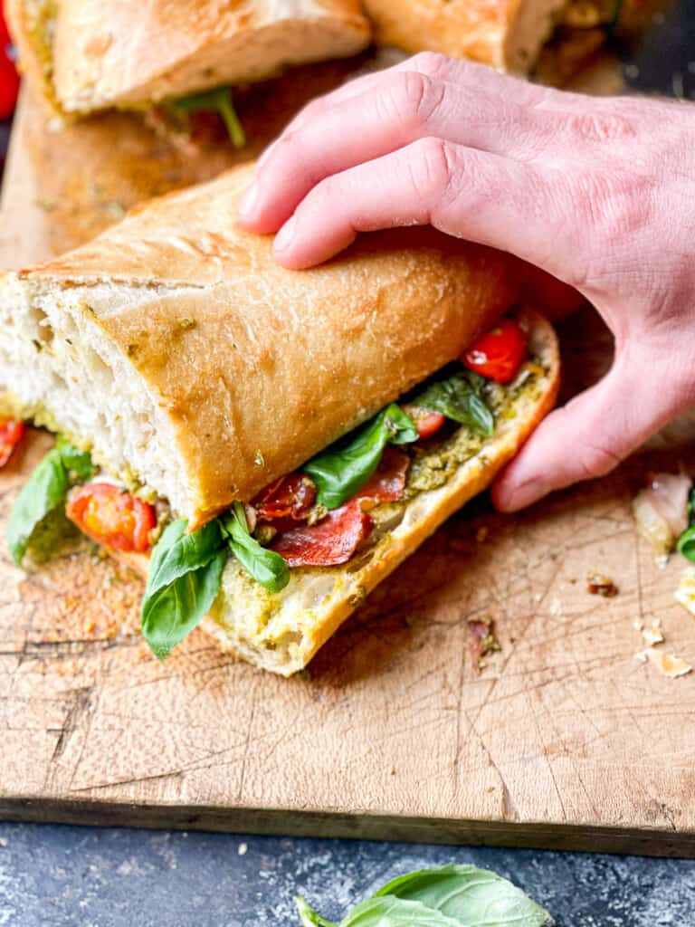 Toasted Caprese Sandwich with Basil Pesto, crispy prosciutto, on a wooden cutting board with a side of tomatoes on the vine for visual appeal. 