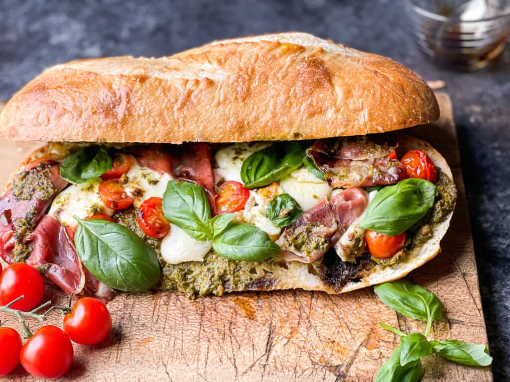 Toasted Caprese Sandwich with Basil Pesto, crispy prosciutto, on a wooden cutting board with a side of tomatoes on the vine for visual appeal. 