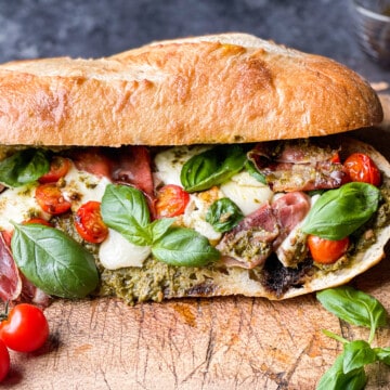 Toasted Caprese Sandwich with Basil Pesto, crispy prosciutto, on a wooden cutting board with a side of tomatoes on the vine for visual appeal.